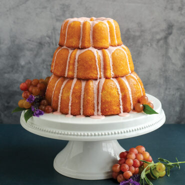 3 tiered baked cakes with glaze on pedestal, grapes around cake
