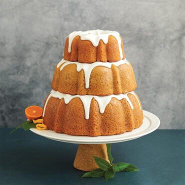 3 tiered baked cakes with glaze on pedestal, half of an orange and some leaves around cake