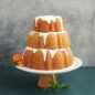 3 tiered baked cakes with glaze on pedestal, half of an orange and some leaves around cake