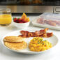 Plated pancakes, bacon, & eggs, bowl of fruit, and glass of orange juice, closed bacon tray in front of microwave