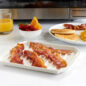 Bacon Tray with bacon strips, plate of pancakes, bacon & eggs, bowl of fruit, glass of orange juice in front of microwave