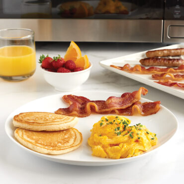 Plate of Eggs, pancakes, & bacon, glass of orange juice, bowl of fruit, and bacon tray with bacon in front of microwave
