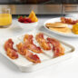 Bacon tray with bacon strips, glass of orange juice, bowl of fruit, plate of pancakes, eggs, & bacon, in front of microwave