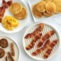 Overhead shot of ribbed and flat side of meat grill with bacon and sausage, plate of eggs, pancakes and bacon, with another plate of pancakes