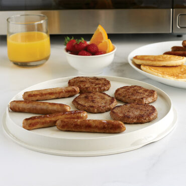 Flat side of meat grill with sausage patties and links, plate of pancakes, bowl of fruit, glass of orange juice, in front of a microwave