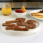 Flat side of meat grill with sausage patties and links, plate of pancakes, bowl of fruit, glass of orange juice, in front of a microwave