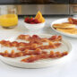 Ribbed side of meat grill with bacon strips, plate of pancakes, bowl of fruit, glass of orange juice, in front of a microwave