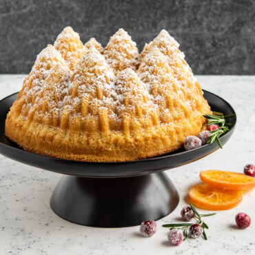 Pine Forest Bundt® cake on stand, covered in powdered sugar, with an orange slice and some berries