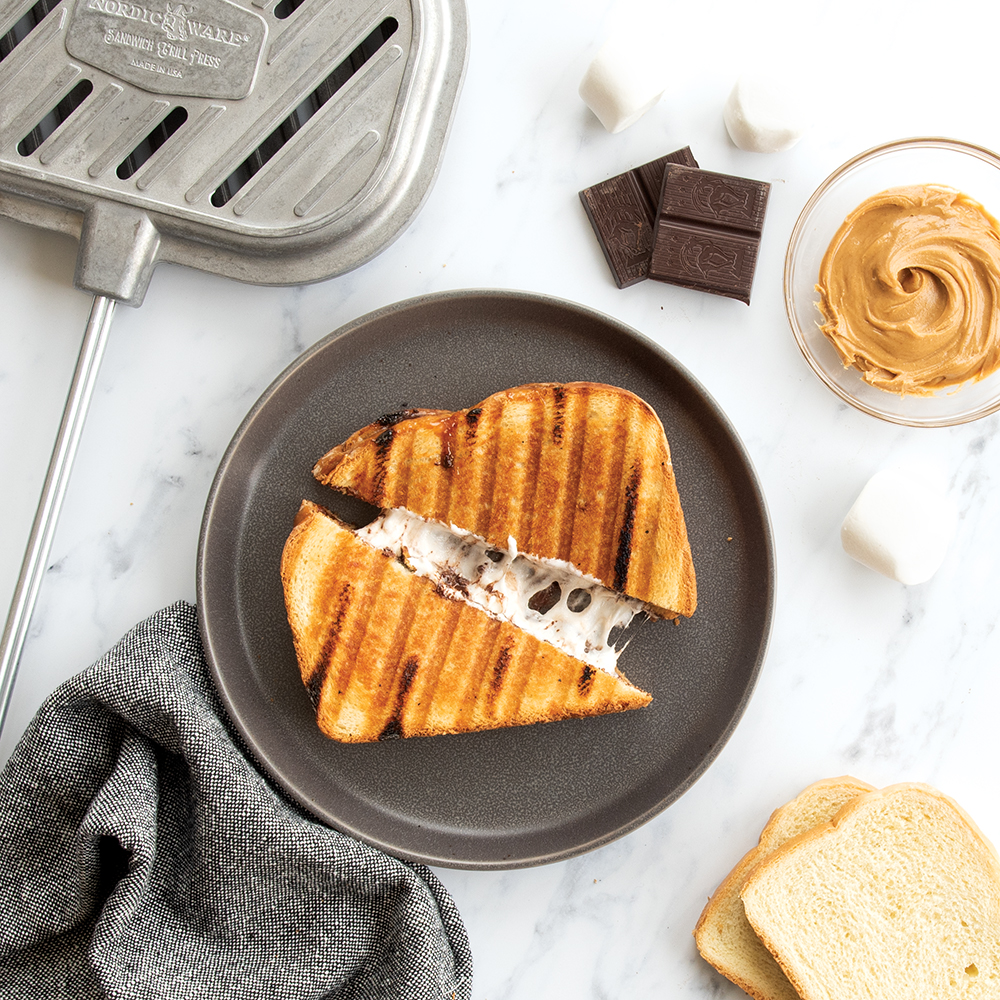 Peanut Butter Smores Sandwich cut in a diagonal on a gray plate. The sandwich is slightly pulled apart to reveal a melted marshmallow center. Next to the plate, there is two slices of bread, a bowl of peanut butter, two marshmellows, two pieces of dark chocolate, a grey towel, and the Campfire Griller. 