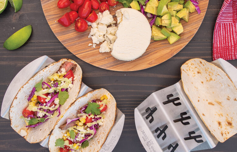 Two Stainless-Steel Taco Grilling Racks with grilled soft tortillas. There is one Taco Grilling Rack on the right with one empty taco shell, and one Taco Grilling Rack on the left with two steak tacos topped with corn, cabbage, cheese, tomatoes, and cilantro. Next to the racks, there is a wooden board with sliced avocado, crumbled queso fresco, chopped tomatoes,, and pieces of cabbages. Next to the board, there are slices of lime and a red kitchen towel. 