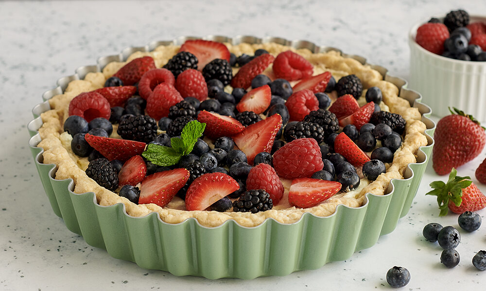 The Aurora Green Quiche and Tart Pan frilled with a mixed berry tart with a vanilla custard filling. The tart is surrounded by raspberries, blueberries, and strawberries. 