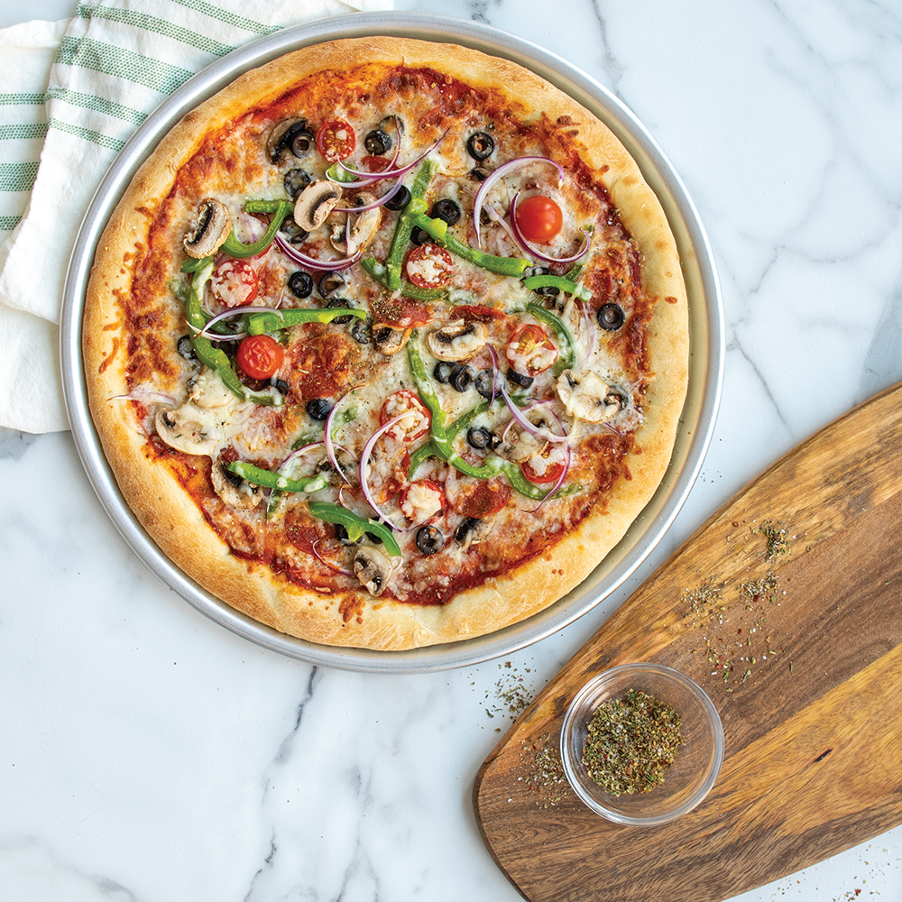 A whole supreme pizza baked in the Naturals Traditional Pizza Pan. This pizza is topped with green peppers, tomatoes, onions, black olives, pepperoni, cheese, mushrooms, and oregano. Next to the pizza, there is a small cup of oregano that was used for sprinkling on top on the pizza. 