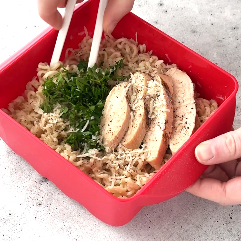 An image of the garlic parmesan noodle recipe being mixed with chopsticks in the Ramen Rush noodle bowl. The noodles are topped with parmesan cheese, herbs, and grilled chicken breast. 