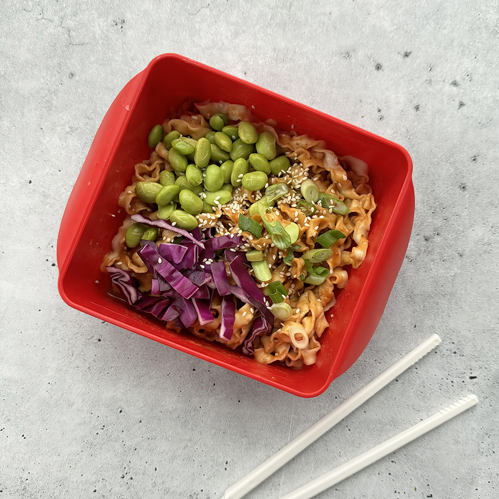 An image of the spicy peanut sesame noodle recipe in the Ramen Rush noodle bowl with Ramen Rush chopsticks on the side. The noodles are mixed with a spicy peanut sesame sauce and topped with edamame beans, purple cabbage, green onions, and sesame seeds. 