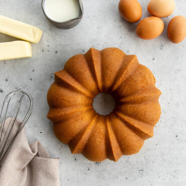 Bundt Cake on countertop surrounded by baking ingredients, sticks of butter, eggs, frosting, egg beater, towel