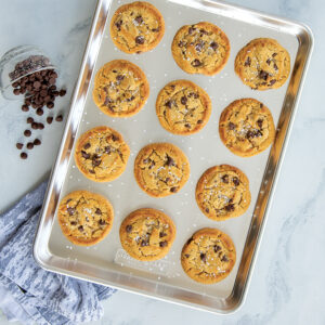 a dozen chocolate chip cookies on a Nordic Ware Naturals Half Sheet next to a bowl of spilled chocolate chips and a blue towel. 