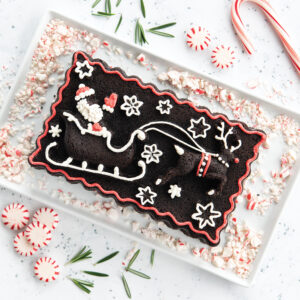 Chocolate Peppermint Loaf Cake made in a Santa Sleigh Loaf Pan decorated with red and white icing. The pan features a detailed design of St. Nicholas and his reindeer. The cake is sitting on a white, rectangle plate with peppermints and rosemary around it. 