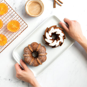 Classic Bundt Duet Cakes on plate being held, with bowl of cinnamon and cooling rack with orange slices